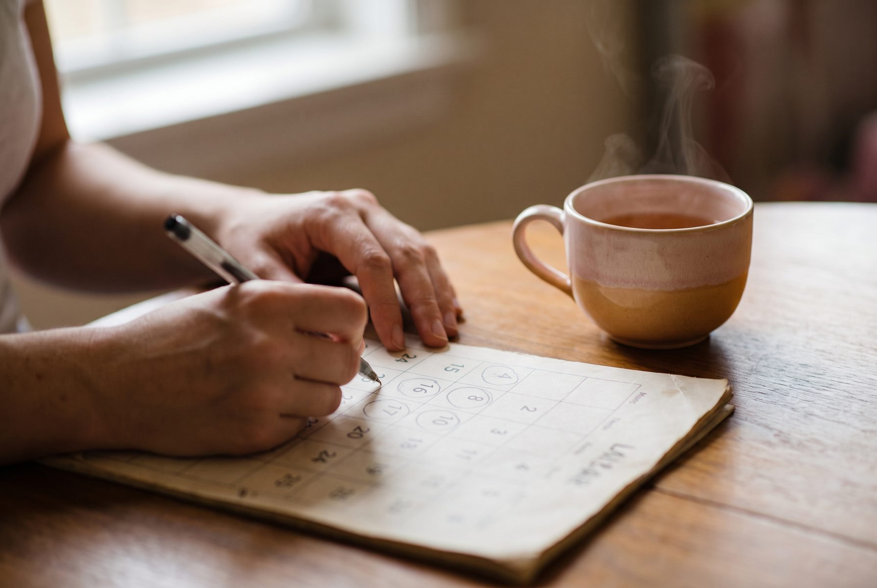 Close-up of a woman's hands marking dates on a paper calendar with a pen, tea cup beside, morning light
