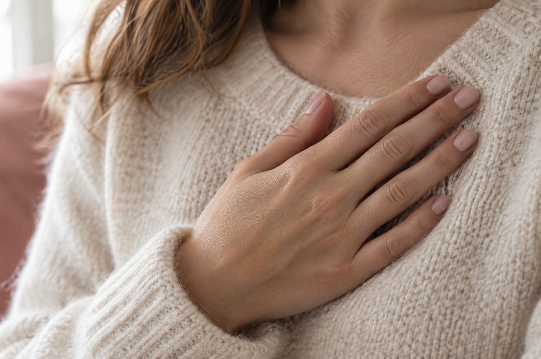 Close-up of a woman's hand resting over her chest on a cream linen sweater, calm soft daylight, no face visible