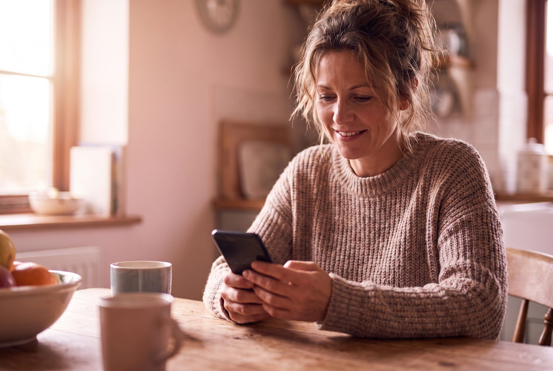 A woman in her mid-40s smiling softly while looking at her phone at a kitchen table, screen not readable