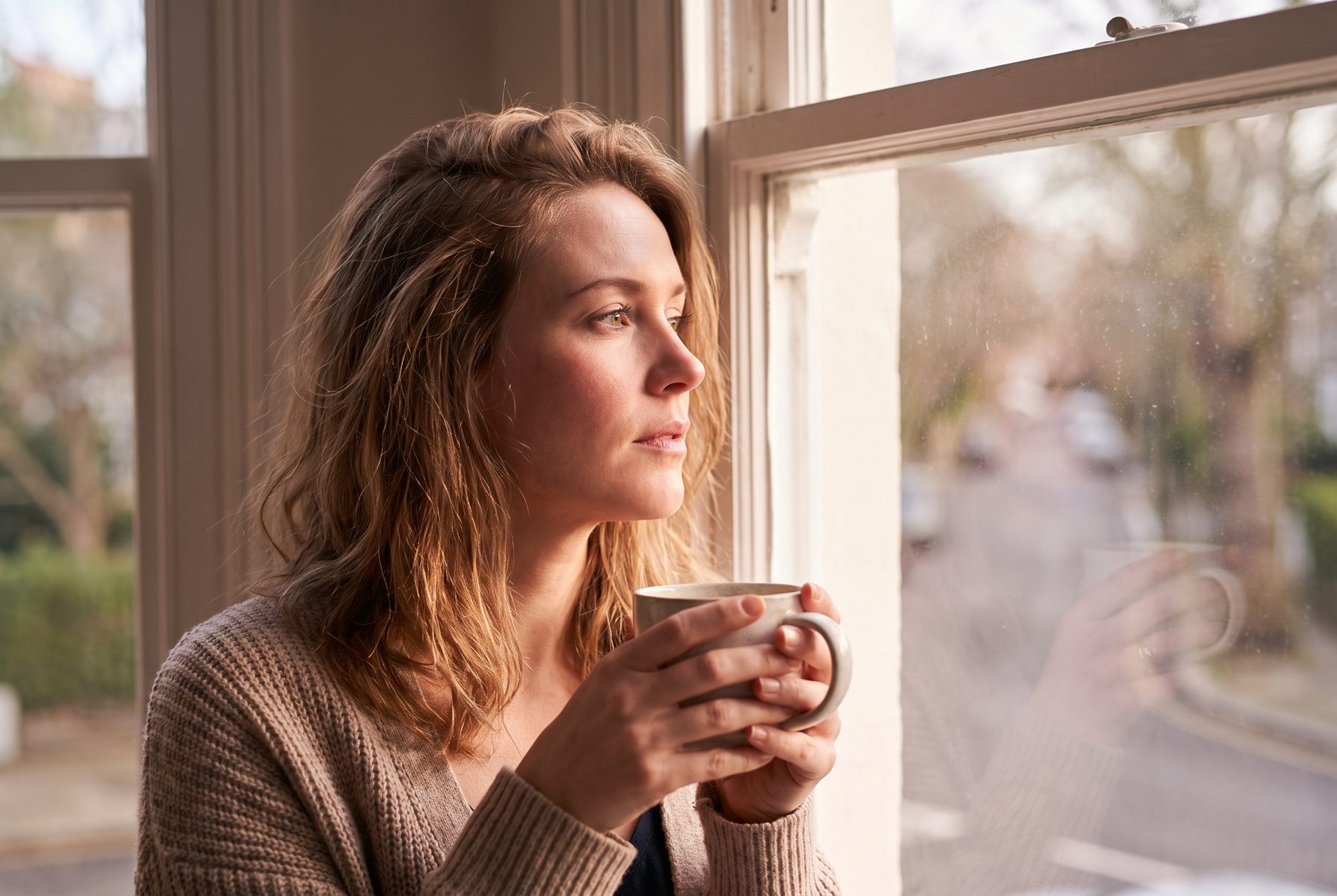 A woman in her mid-40s standing at a bright window with a cup of tea, thoughtful expression, American look