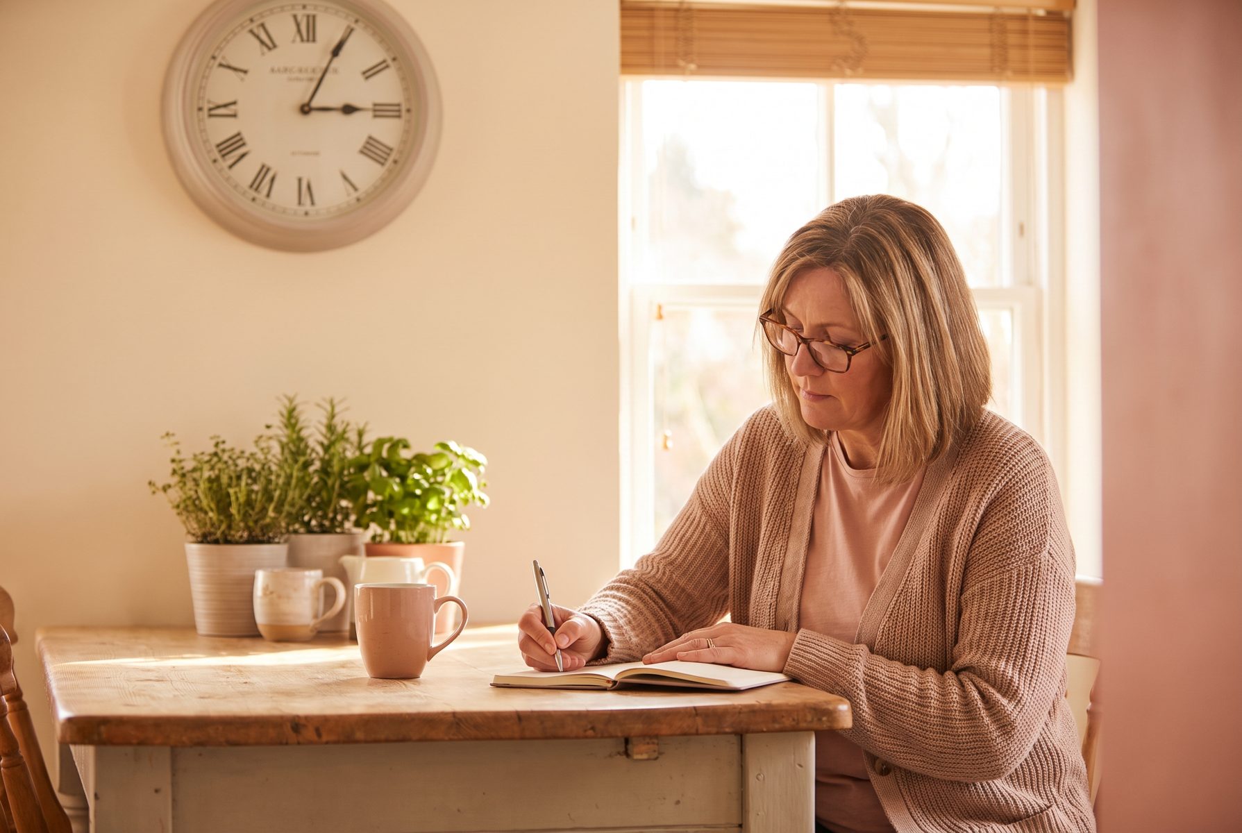 A woman in her late 40s writing notes at a kitchen table with a wall clock behind her, focused, morning light